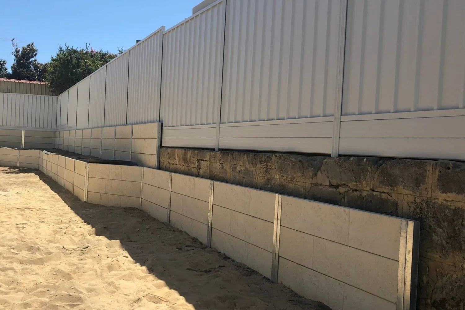 Tiered white concrete sleeper retaining walls on a sandy block below a Colorbond fence